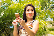 日本中年妇女在热带花园看远了手机在手里