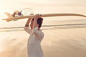 一个夏天的晚上，在康沃尔的Gwithian Towans海滩，一个女人背着一个冲浪板在日落附近。