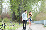 一个大学男生帮他的女朋友玩滑板穿过公园