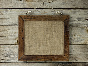 旧风化破旧的天然木相框与粗麻布袋材料框在里面，挂在一个钩子上的旧风化木面板墙的背景。