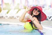 美丽的年轻女子穿着比基尼在游泳池与太阳帽和鸡尾酒