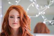 14 / 15岁的红发少女站在墙上的镜子前看着镜头，被仙女般的灯光照亮，姜黄色的卷发，自然的妆容，苍白的皮肤和雀斑