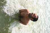 图片是印度男子在海滩度假，在大海/印度洋中游泳和戏水，在印度南部果阿/喀拉拉邦海滩的海边背景下，向一侧看，大笑，看起来很开心