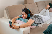 一位成年女性躺在客厅沙发上的垫子上，使用耳机和智能手机