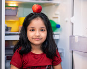 可爱的小女孩拿着苹果站在冰箱旁边。