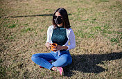 戴着面具和太阳镜的年轻女子盘腿坐在草地上，手里拿着咖啡。