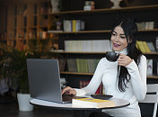年轻漂亮的女人喝咖啡在黑马克杯看在咖啡馆的笔记本电脑
