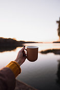女人的手握着一杯茶在湖的背景在日落