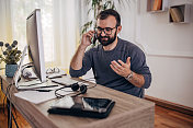 男人在家里办公室的电脑上工作，用智能手机说话