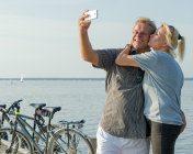 微笑的老年夫妇在阳光下拍摄自拍照在码头上的湖在自行车后休息