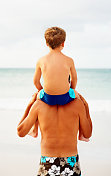 一个父亲背着他的儿子在海滩上的后视图