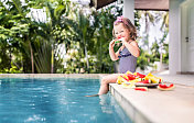可爱的女孩坐在游泳池边吃西瓜